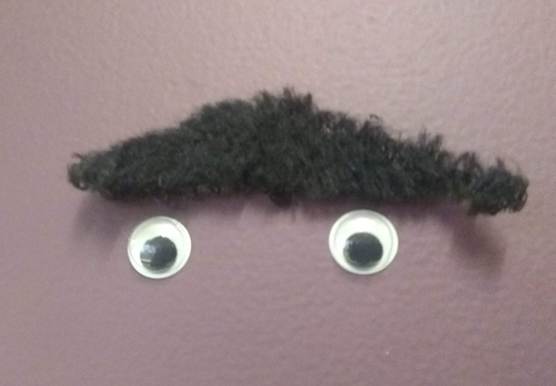 Googly eyes stuck to a wall below a fake moustache.