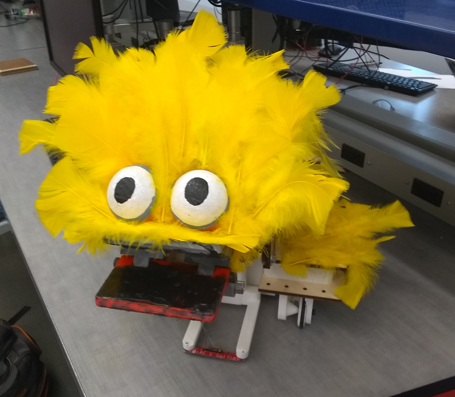 Googly eyes on a wheeled robot with a grabber, decorated to look like Big Bird with craft feathers.