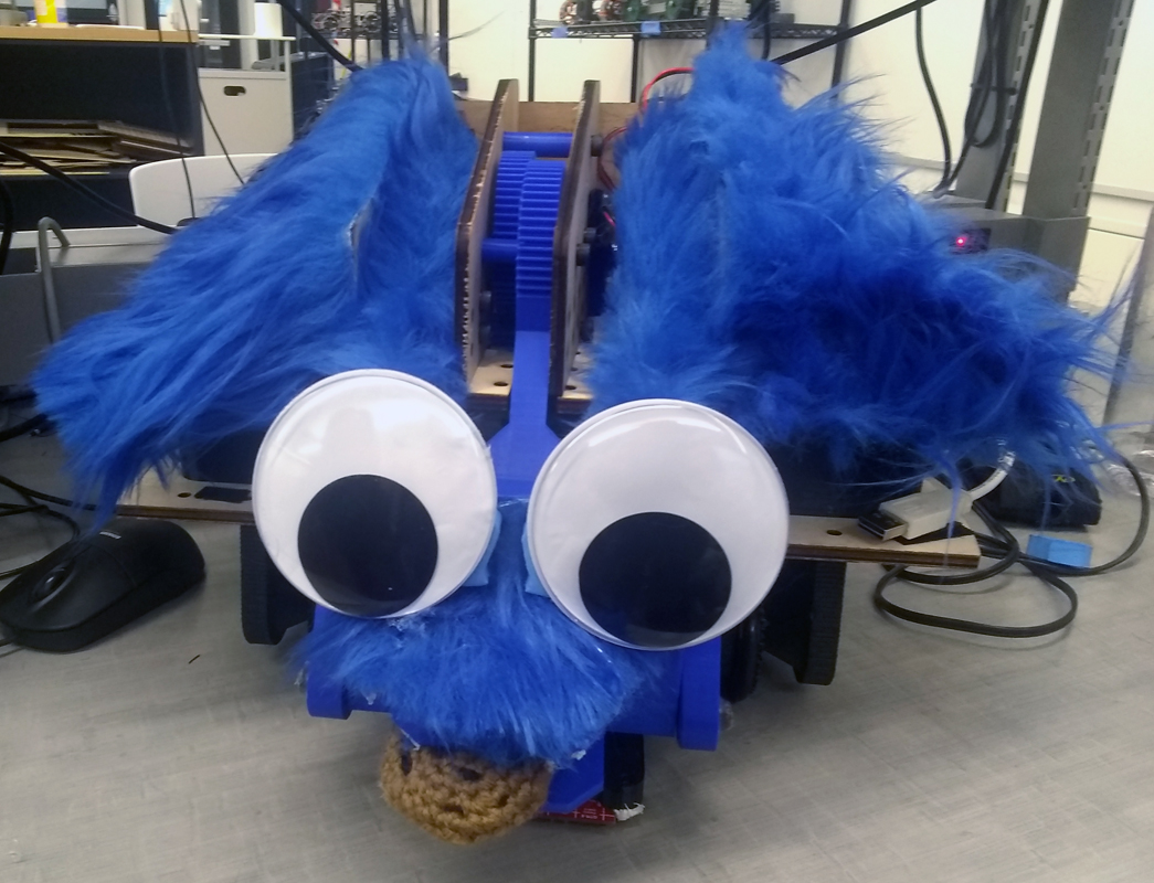 Googly eyes on a wheeled robot with a grabber, decorated to look like cookie monster with craft feathers.
