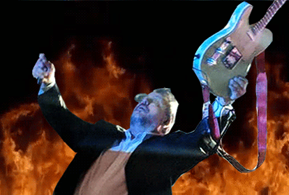John F raises his guitar as flames rise behind him.
